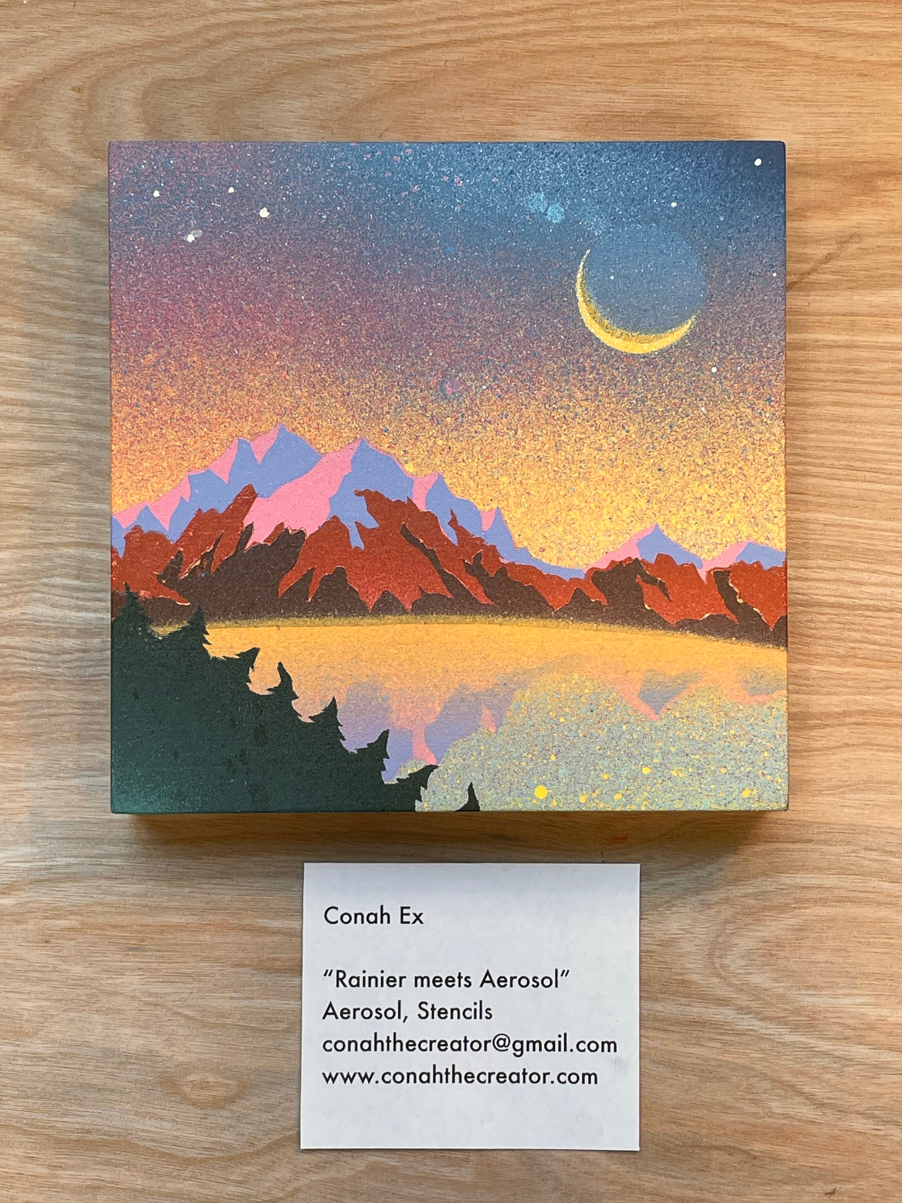 Artistic depiction of a mountain landscape with a crescent moon on a wooden surface, featuring a business card below.