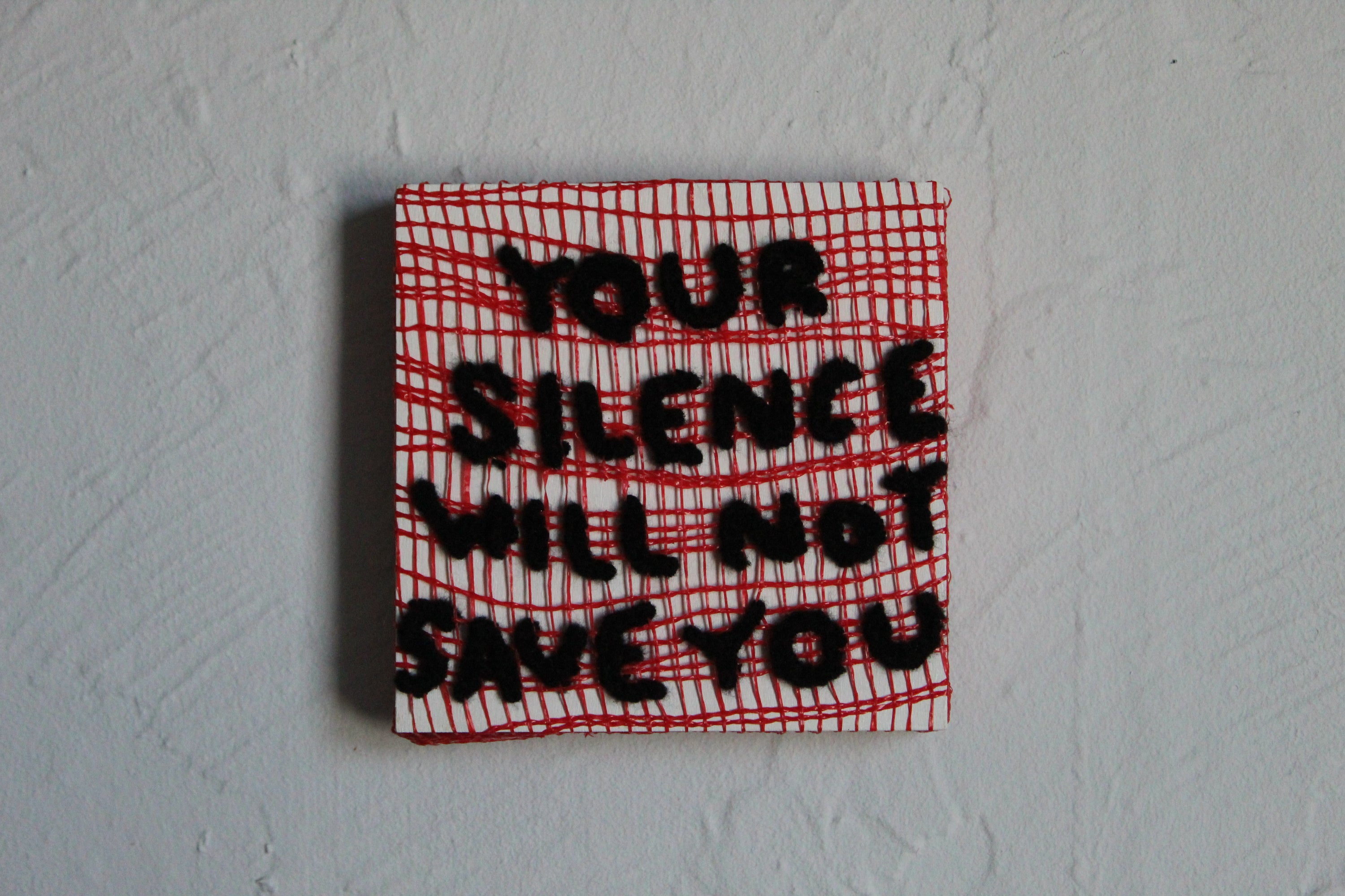Textile art piece with 'Your silence will not save you' on a red and white checkered background.