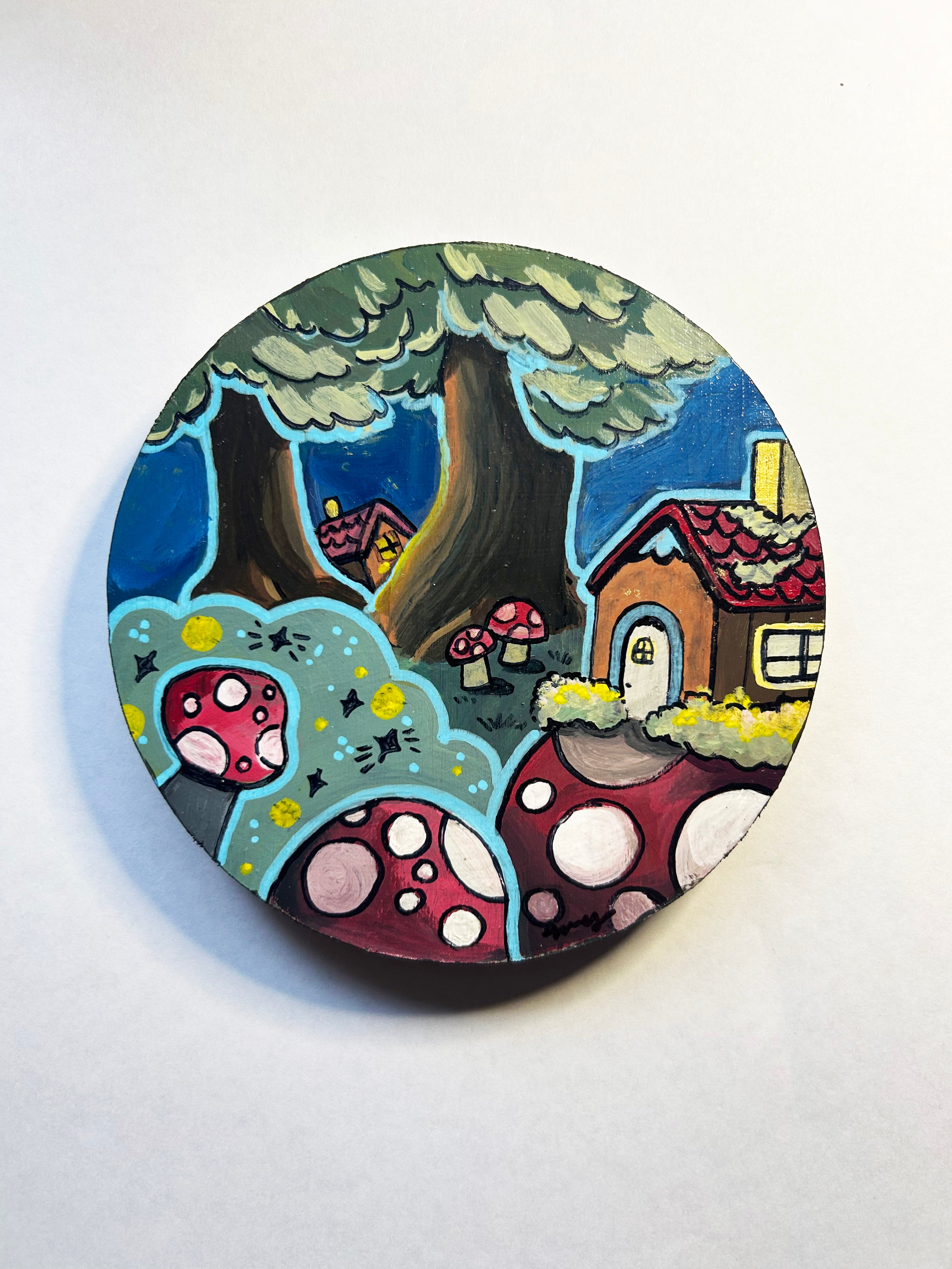 Round painted coaster with a whimsical forest design featuring a house, trees, and mushrooms on a white background.