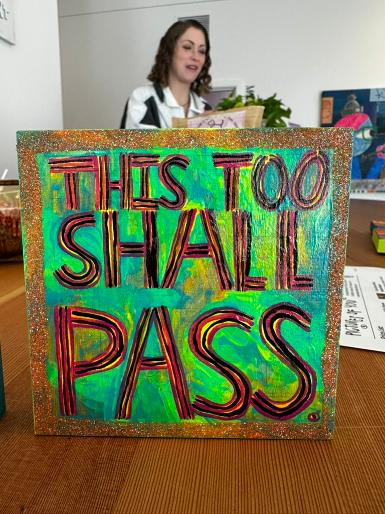 Colorful sign with 'This Too Shall Pass' text in an office setting