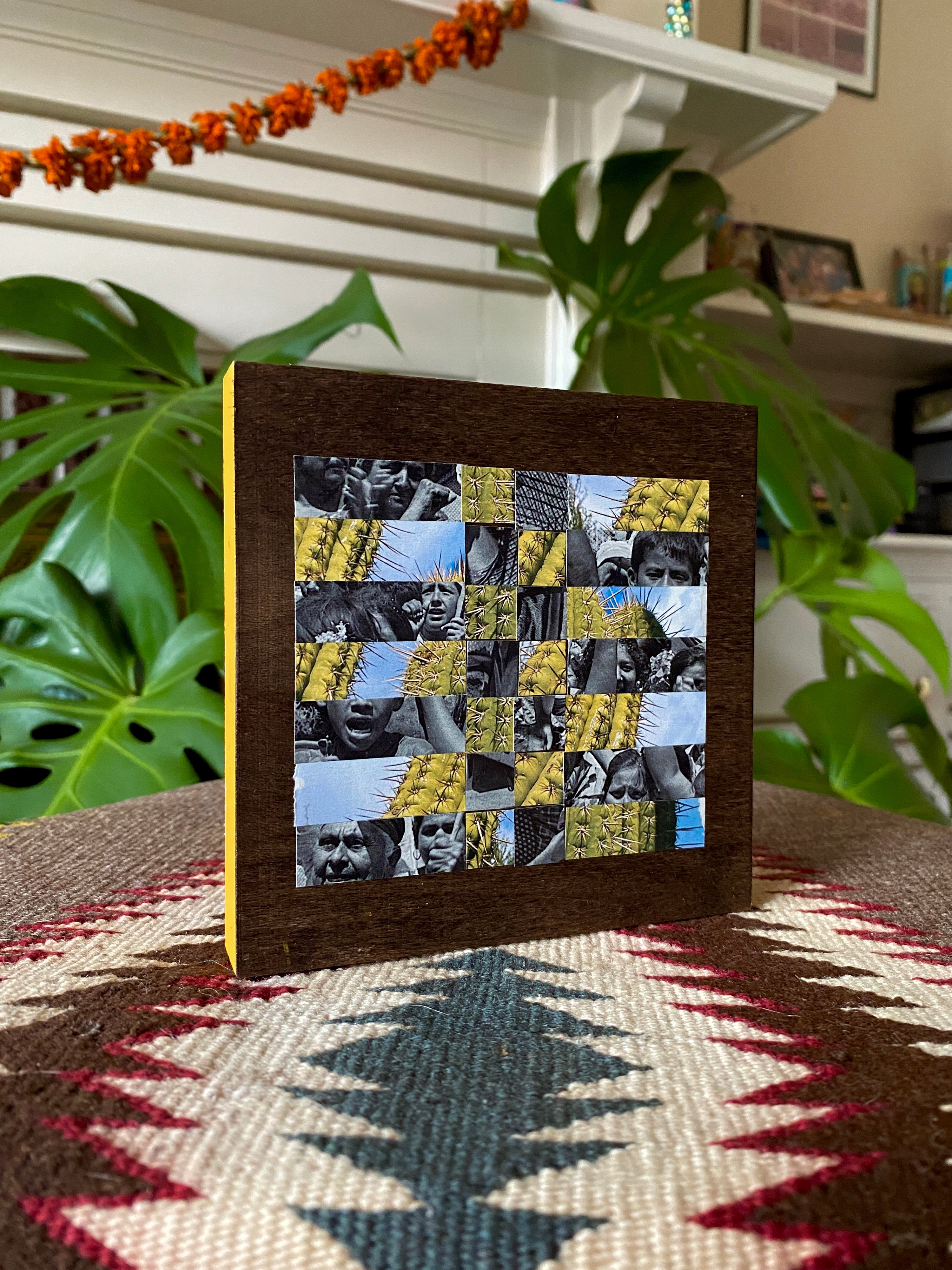 Framed mosaic art piece on a table with plants and a fireplace in the background